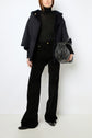 A person wearing a black fur-lined jacket reminiscent of a manteau en mouton, paired with a black turtleneck and patterned black pants, holds a black handbag. They are standing against a plain white background.