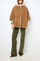 Woman wearing a tan cape peau lainée with a hood, green patterned pants, brown shoes, and sunglasses, standing against a plain white background.