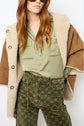 A woman wears sunglasses, an olive shirt, patterned green pants with a chain belt, and a manteau cuir femme draped over her shoulders.