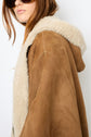 A person with long brown hair and sunglasses wears a stylish manteau cuir femme—a brown suede coat with a cream shearling lining and hood—shown from the side against a white background.