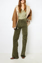 A woman wearing a tan shearling jacket reminiscent of a classic manteau cuir femme, green button-up shirt, olive patterned pants with a chain belt, and brown shoes stands against a white background.