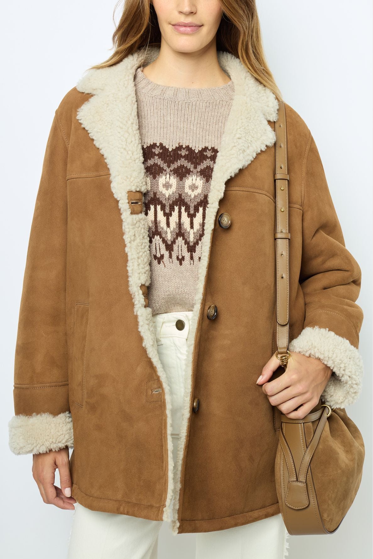 A person wears a brown shearling coat, reminiscent of a manteau mouton velours, over a beige patterned sweater, paired with white pants and carrying a brown shoulder bag. The vintage-inspired coat features fluffy white lining on the collar, cuffs, and edges.