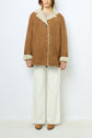 A person wearing a manteau vintage brown shearling coat, cream sweater, white wide-leg pants, and brown shoes stands against a plain white background.