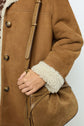 A person wearing a brown shearling coat, reminiscent of a manteau mouton velours, holds a tan suede and leather shoulder bag. Only their torso and hand are visible.