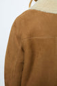 Close-up of a person wearing a brown suede jacket with a cream-colored shearling collar, shown from the back. Only part of the shoulder and upper arm is visible, highlighting the timeless appeal of this manteau mouton velours.