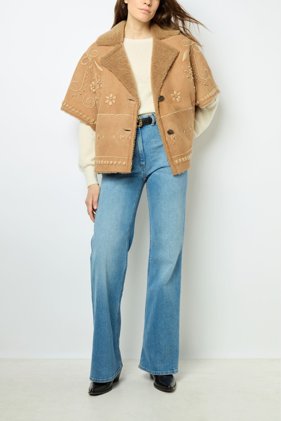 A woman wearing a veste courte oversize peau lainée embroidered with fleurs brodées over a cream sweater, blue high-waisted jeans, and black shoes, standing against a plain white background.