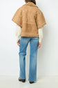 Person wearing light blue jeans, a cream sweater, and a tan peau lainée cape with fleurs brodées, standing against a white background, facing away from the camera.