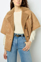 A woman wearing a tan peau lainée shearling jacket with fleurs brodées over a cream sweater and blue jeans, standing against a light background.