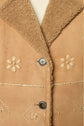 Close-up of a tan peau lainée shearling coat with a fuzzy collar, fleurs brodées flower-shaped embroidery, and two visible buttons. A white knit garment is partially visible underneath.