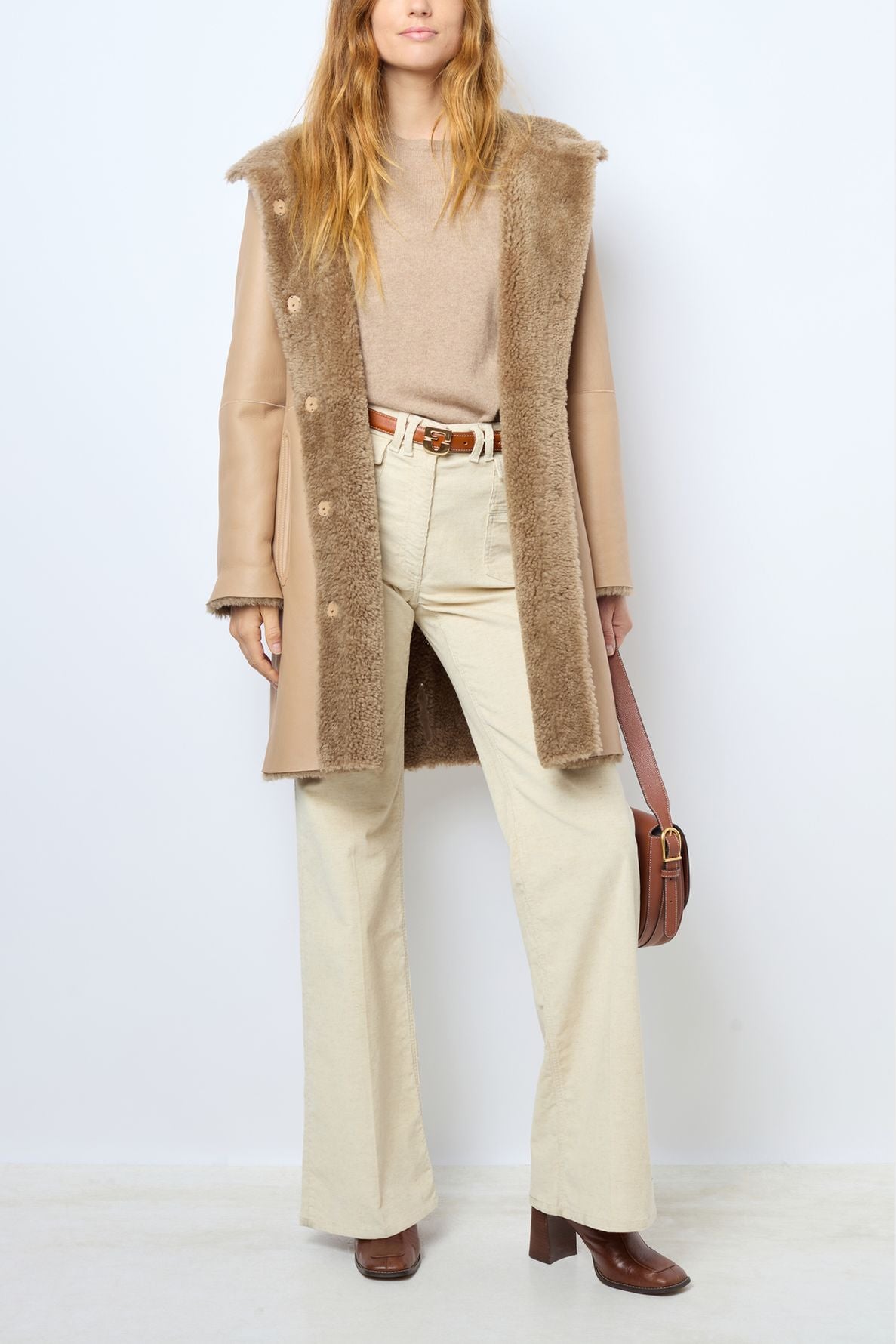 Woman wearing a tan manteau peau lainée, beige sweater, cream pants, brown heeled shoes, and holding a brown purse. Outfit is neutral and stylish, with a mix of textures. Background is plain white.