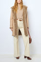 Woman wearing a tan manteau peau lainée, beige sweater, cream pants, brown heeled shoes, and holding a brown purse. Outfit is neutral and stylish, with a mix of textures. Background is plain white.
