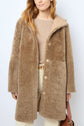 A woman wearing a light brown manteau réversible faux shearling coat over a beige sweater and cream pants, holding a brown handbag, stands against a plain white background.