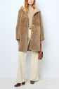 A person wearing a chic manteau peau lainée in tan faux fur, paired with a beige sweater, cream wide-leg pants, brown boots, and carrying a brown shoulder bag stands against a plain light-colored background.