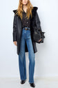 A woman wearing a black cuir d’agneau faux fur-lined coat, black polka dot blouse, high-waisted blue jeans, and black boots, holding a black handbag, stands against a plain white wall.