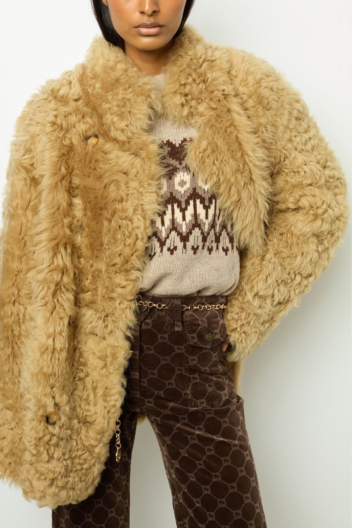 A person wearing a fluffy tan manteau court faux fur coat, a beige sweater with a brown geometric pattern, and brown patterned pants with a chain belt, standing against a plain white background.