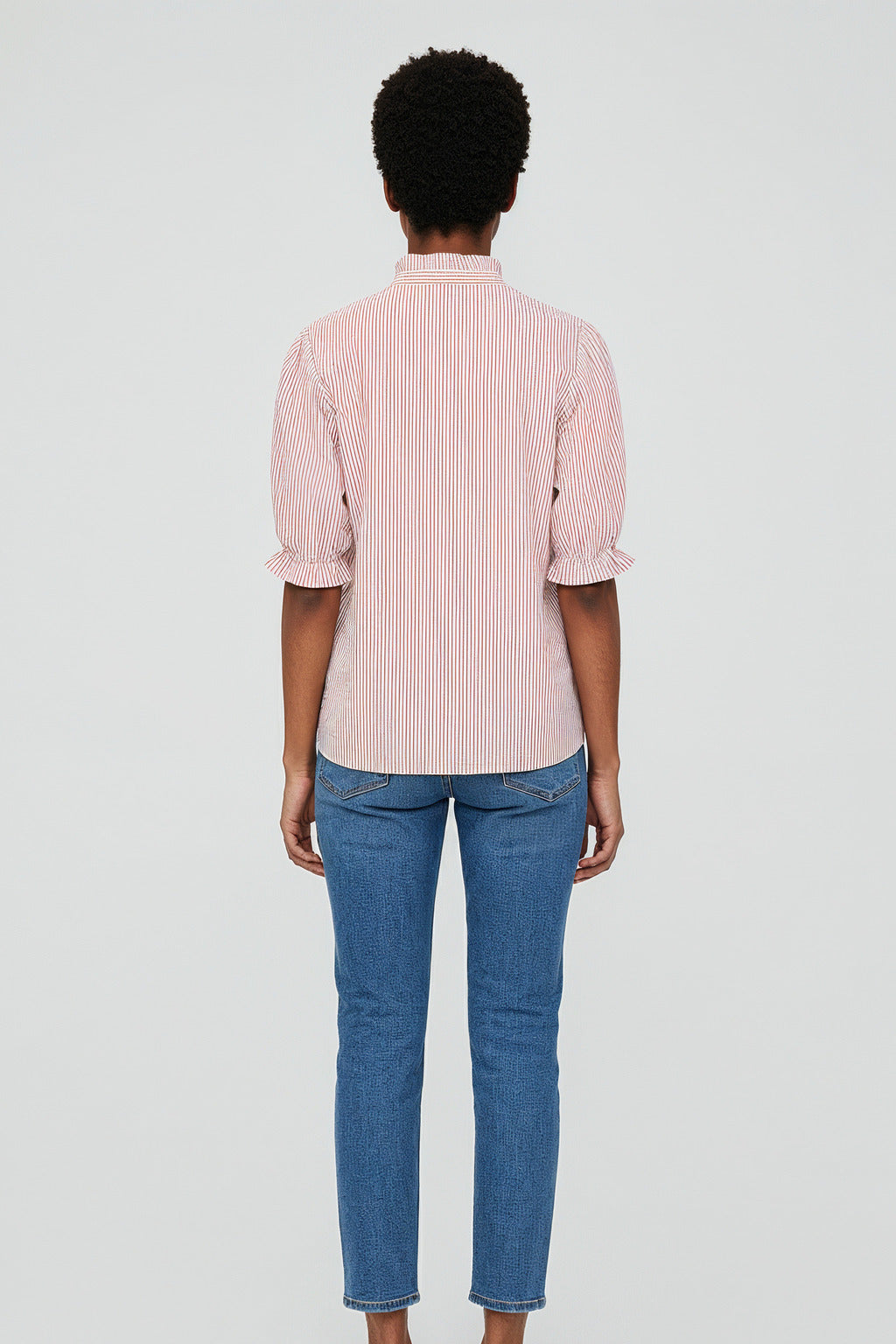 A person stands facing away, wearing a red and white striped blouse en coton with manches bouffantes and blue jeans. The background is plain and light gray.