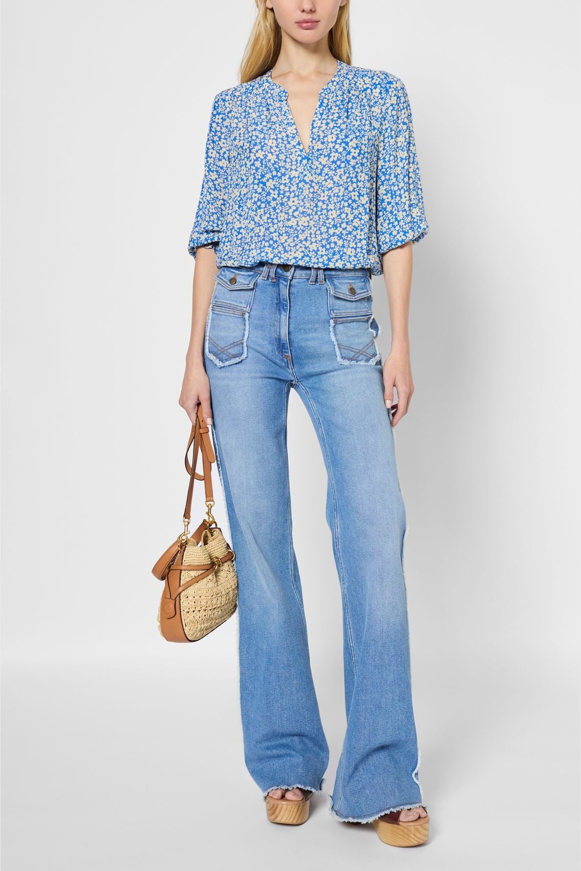 Woman wearing a blue blouse fluide with a motif floral design and manches 7/8, paired with high-waisted flared jeans and holding a tan and straw handbag. She is also wearing tan platform sandals. The background is white.