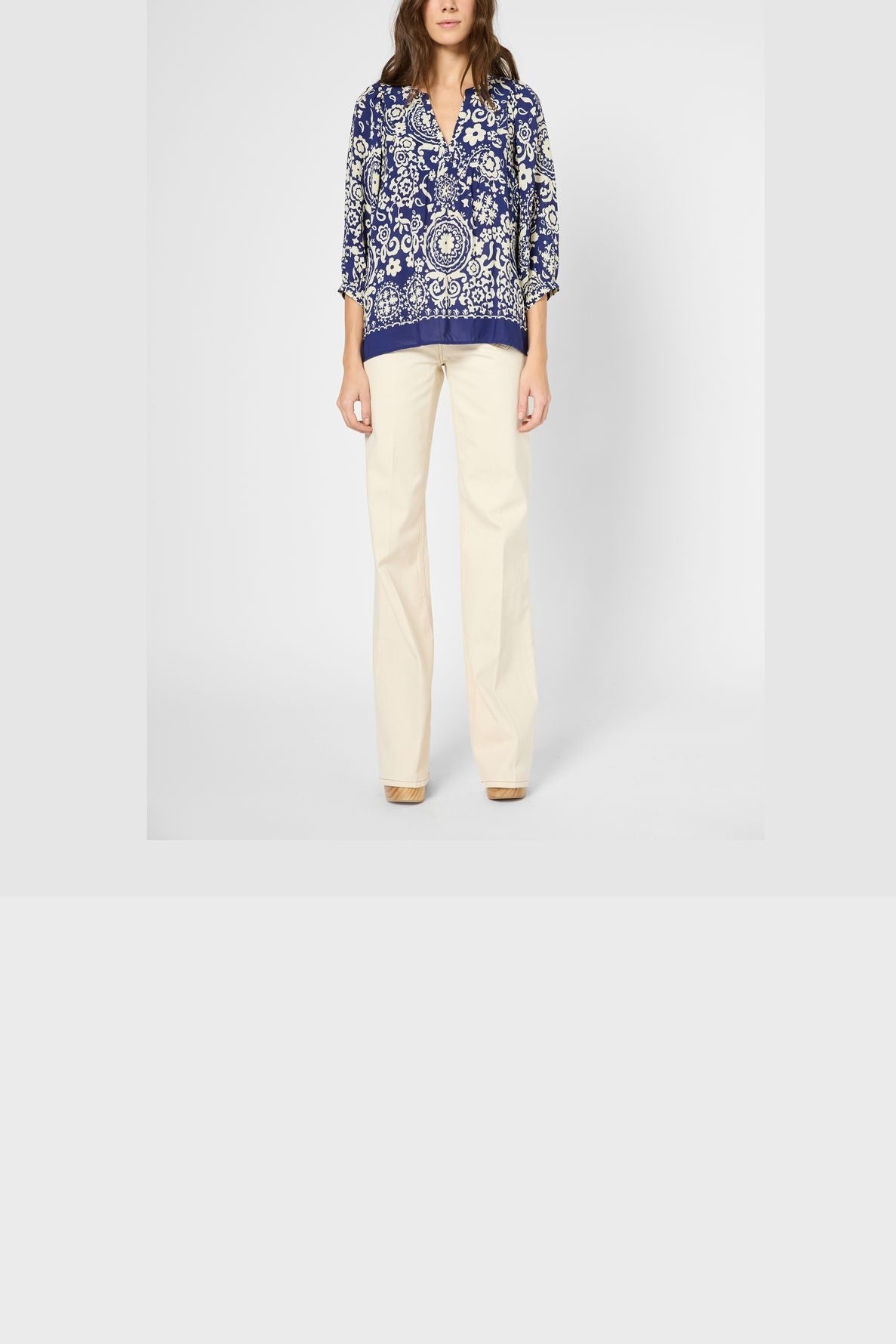 A woman stands facing forward, wearing a blouse fluide with a blue motif floral pattern and three-quarter sleeves, paired with cream-colored wide-leg pants. Her hands are relaxed by her sides against a neutral background.
