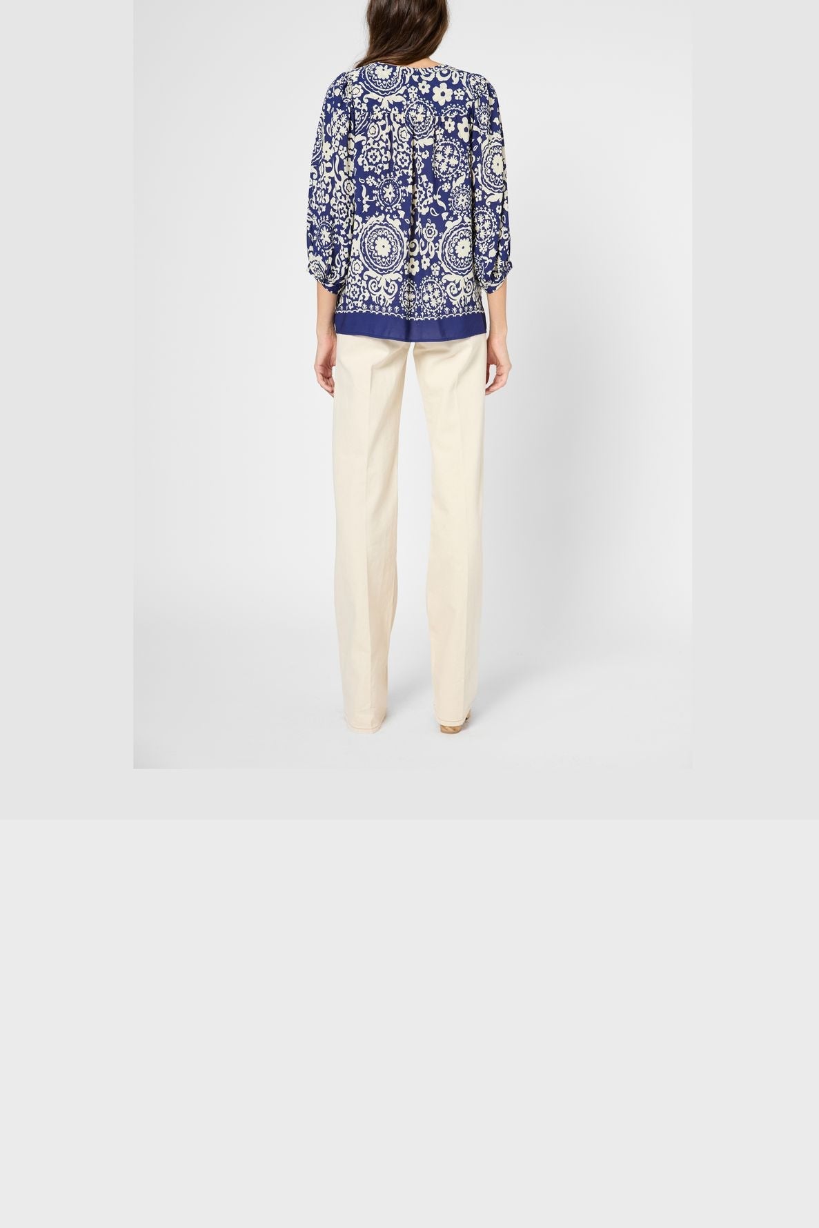 Woman standing with her back to the camera, wearing a blue and white blouse fluide with an imprimé graphique and cream-colored pants against a plain white background.