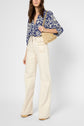 Woman wearing a blouse fluide with an imprimé graphique in blue tones, high-waisted cream wide-leg pants, beige platform shoes, and holding a woven straw handbag. She stands against a plain light background. Her head is slightly cropped from the image.