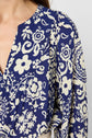 Close-up of a person wearing a blue and white blouse fluide with a floral motif, featuring a V-neck and long sleeves. Only the left side of the upper body and part of the face are visible.
