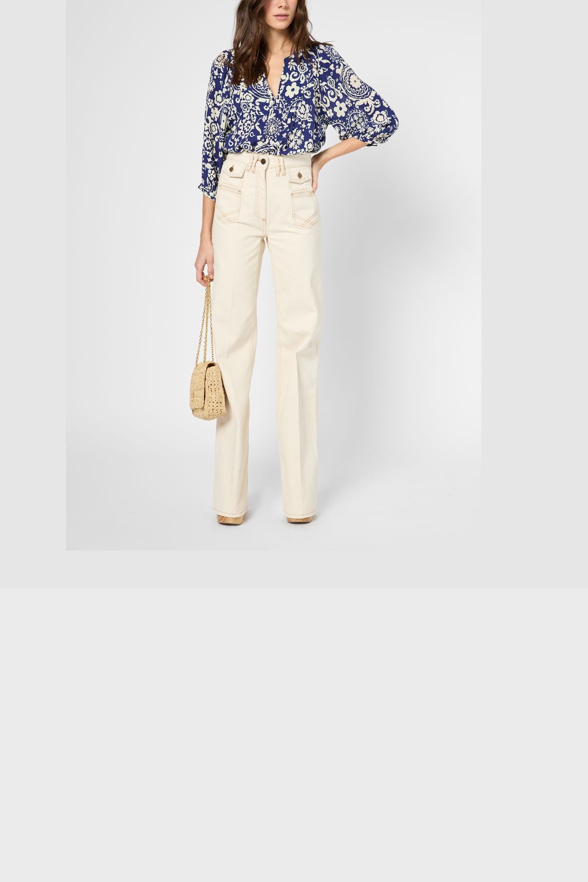 A woman wears a blue blouse fluide à imprimé graphique, high-waisted cream pants, and beige shoes. She holds a small beige woven bag. Her hair is loose and her face is partially out of frame.