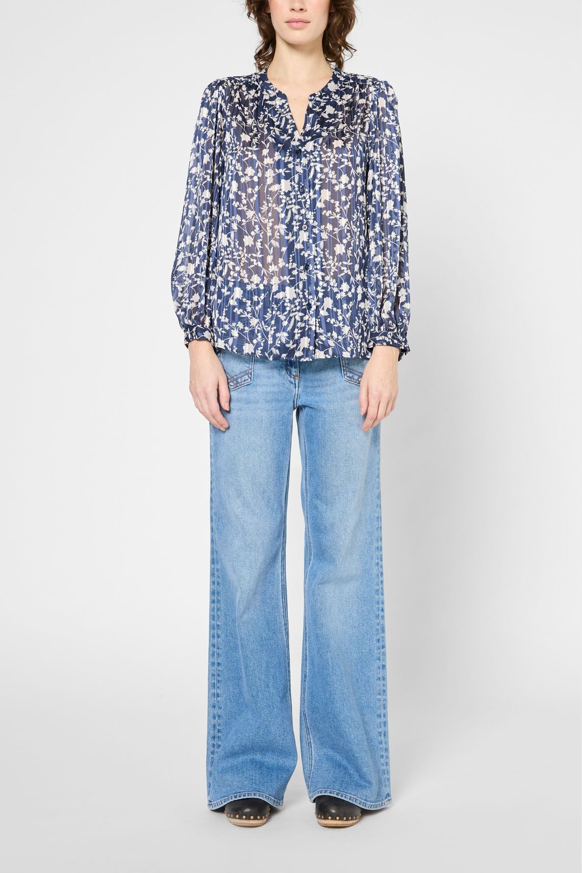 A person wearing a blue and white floral blouse with long sleeves and light blue wide-leg jeans, standing against a plain white background.