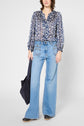 Person wearing a blue floral blouse, light blue wide-leg jeans, and black clogs, holding a black tote bag, standing against a plain white background.
