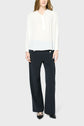 A person wearing a loose white blouse fluide à manches longues, dark wide-leg trousers, and black pointed shoes stands against a plain light gray background. Their face is partially out of frame.