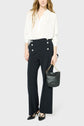 A person wearing a loose white blouse satin with manches longues, high-waisted black pants with front buttons, black heels, and holding a small black handbag with a gold chain strap, stands against a plain light background.