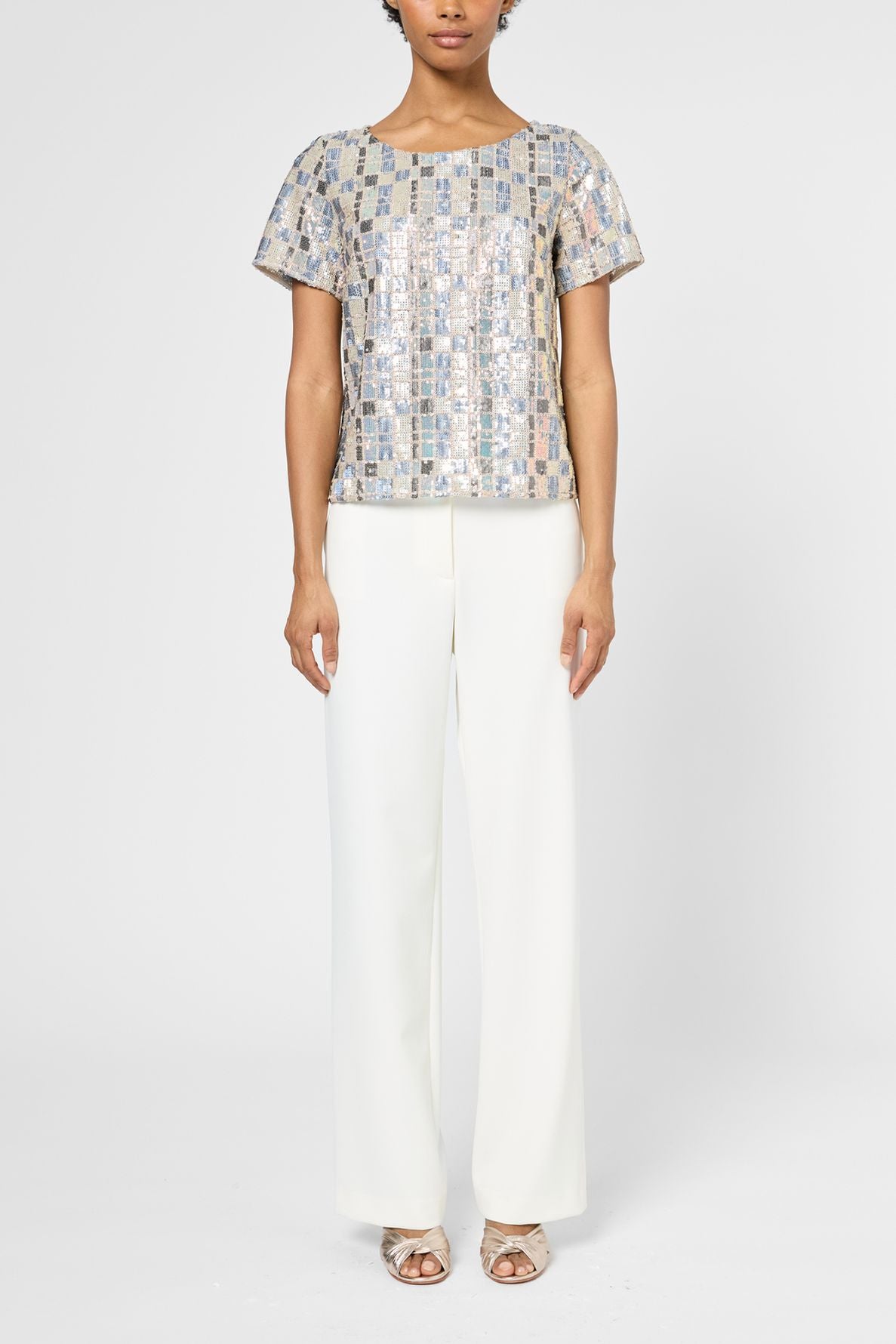 Person wearing a short-sleeve, silver sequin top with white wide-leg pants and open-toed heels, standing against a plain white background.