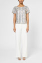 Person wearing a short-sleeve, silver sequin top with white wide-leg pants and open-toed heels, standing against a plain white background.
