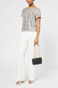A woman in a short-sleeve patterned top, white trousers, and metallic sandals holds a black purse with a gold chain strap. She is standing against a plain light background.