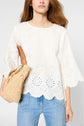 Woman wearing a white eyelet top with scalloped edges and blue jeans, holding a woven straw bag with a brown handle.