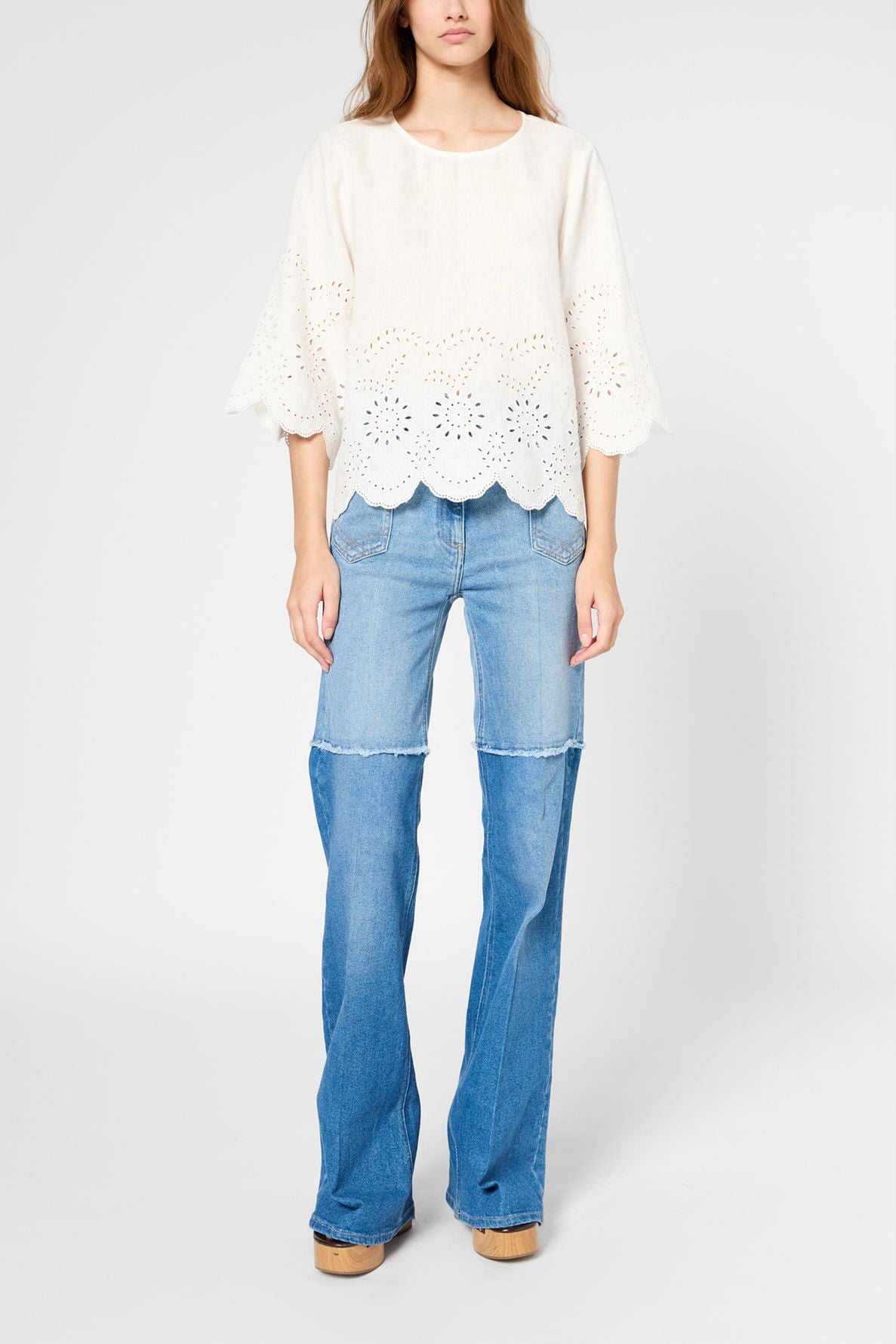 A person wearing a loose, cream eyelet top with scalloped edges and flared blue jeans stands against a plain light background. The face is not visible.