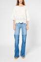 A person wearing a loose, cream eyelet top with scalloped edges and flared blue jeans stands against a plain light background. The face is not visible.