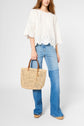 A woman in a white eyelet top and blue flared jeans holds a woven straw handbag with wooden handles. She is standing against a plain light background.