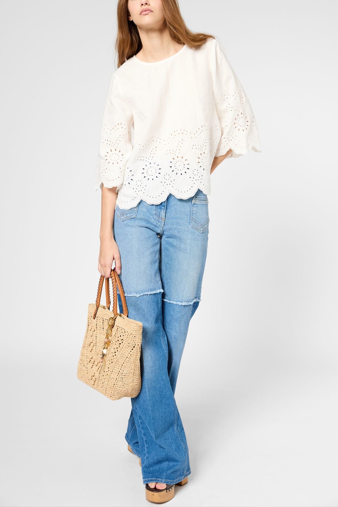A person wearing a white eyelet blouse, blue flared jeans, and platform sandals holds a woven tan bag. Their long brown hair is loose, and their face is partially out of frame.