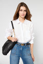 A woman with long brown hair wears a white button-up shirt and high-waisted blue jeans, holding a black shoulder bag with a gold chain strap, standing against a plain light background.