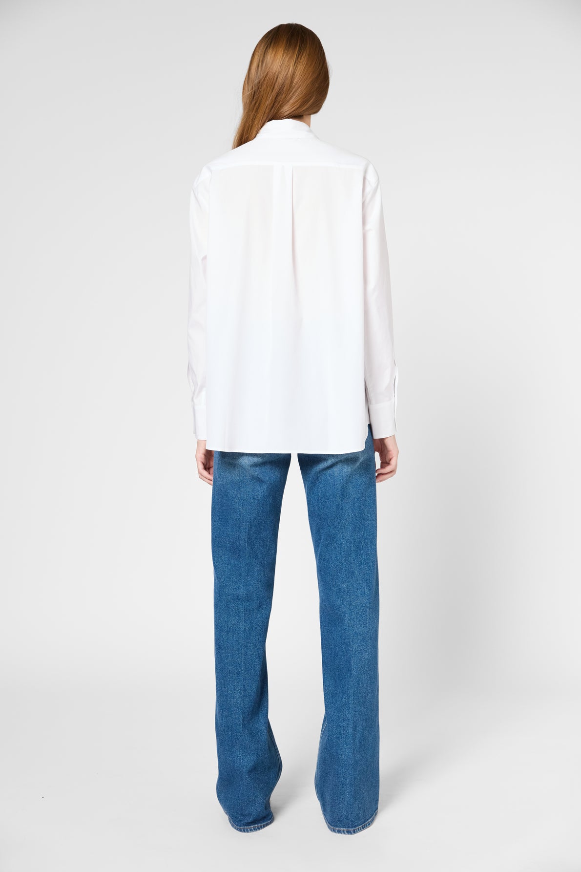 A person with long hair, seen from behind, is wearing a loose white button-up shirt and wide-leg blue jeans, standing against a plain white background.