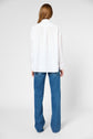 A person with long hair, seen from behind, is wearing a loose white button-up shirt and wide-leg blue jeans, standing against a plain white background.