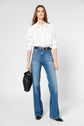 A woman wearing a white button-up shirt, blue high-waisted jeans, and black shoes stands holding a black handbag against a plain light background.