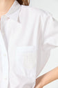 A person wears a white button-up shirt with a chest pocket featuring a decorative lace patch. Only part of the upper body and one arm is visible against a plain background.