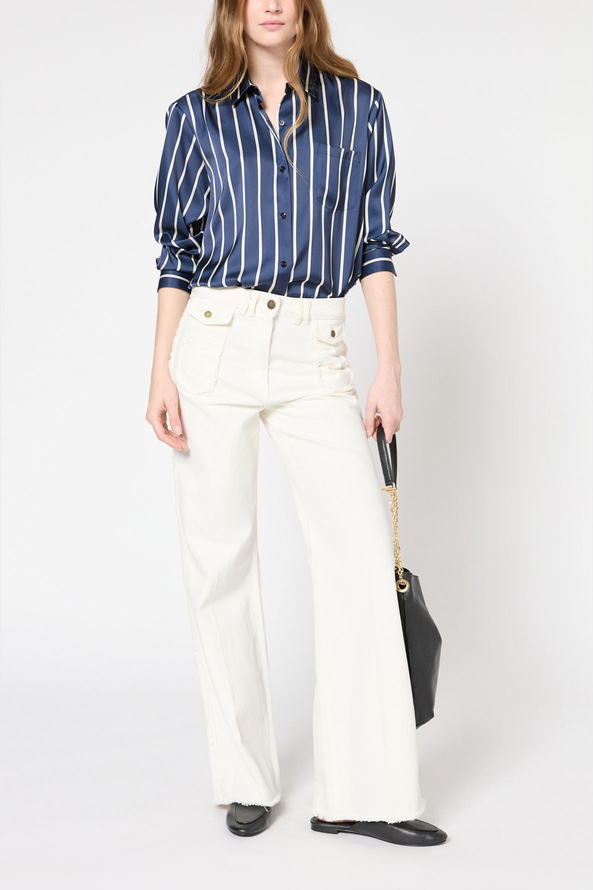 Woman wearing a navy blue and white vertical striped chemise satinée with long sleeves and white high-waisted wide-leg pants, holding a black handbag with a chain strap, and wearing black shoes.
