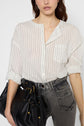 A woman wearing a white, striped button-up shirt tucked into high-waisted gray jeans holds a large black leather handbag. Her sleeves are rolled up and her hair is loose. The background is plain white.