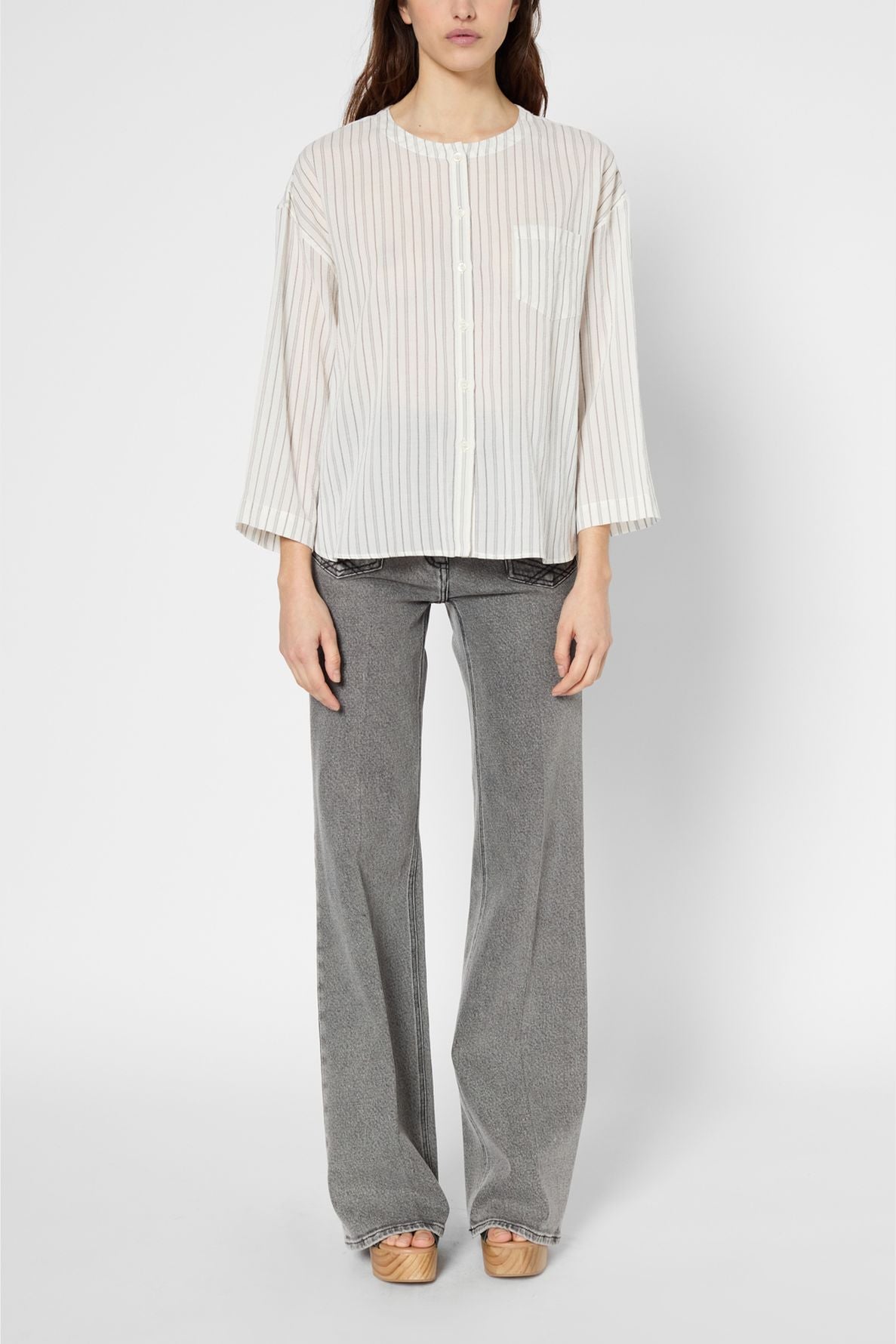 A person wearing a sheer, white, long-sleeve, striped button-up shirt with gray wide-leg jeans and beige shoes, standing against a plain white background.