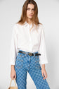 A woman wearing a white button-up shirt and blue patterned jeans with a belt stands against a plain background, holding a straw bag.