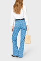 A person with long, wavy hair wears a white shirt, patterned blue jeans, black shoes, and holds a woven beige handbag. The person is facing away from the camera against a plain light background.