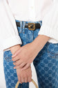 A person wearing a white long-sleeve shirt, blue patterned jeans, and a black belt with a gold buckle stands with hands crossed, holding a rope handle.