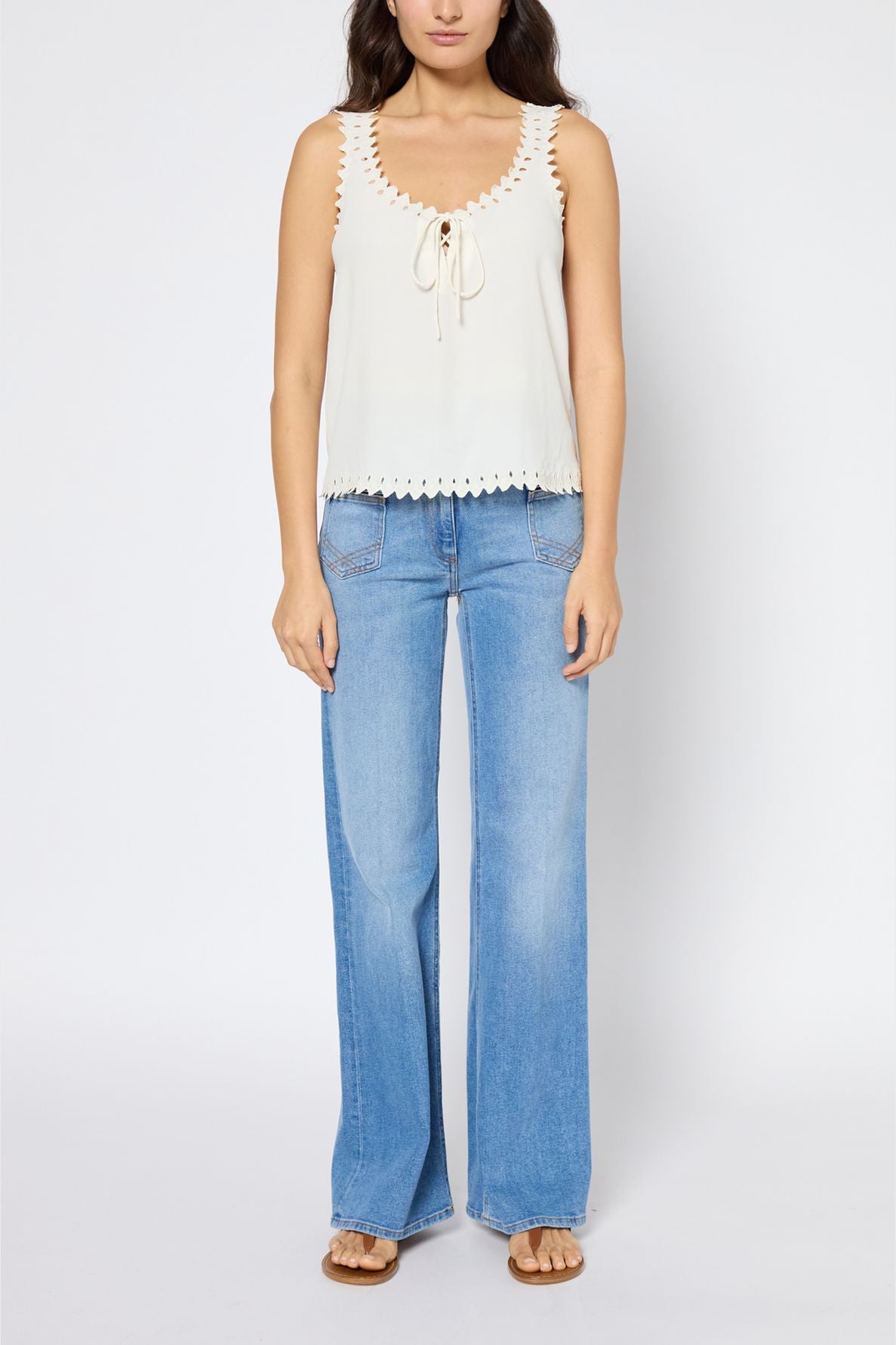 Woman wearing a sleeveless cream top with scalloped edges, blue wide-leg jeans, and sandals, standing against a plain white background.